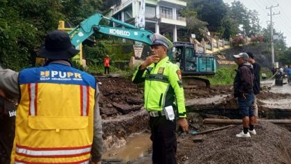 Seorang polisi lalu lintas dan petugas PUPR mengawasi proses perbaikan jalan amblas di Jalan Poros Enrekang-Toraja, dengan alat berat ekskavator bekerja di latar belakang.